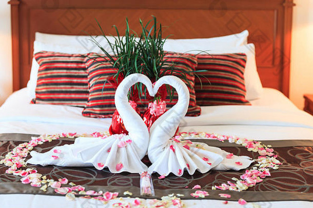 浪漫的卧室内部，亲吻天鹅的折纸毛巾和洒在床上的新鲜粉红白色玫瑰花瓣装饰新婚夫妇。威丁