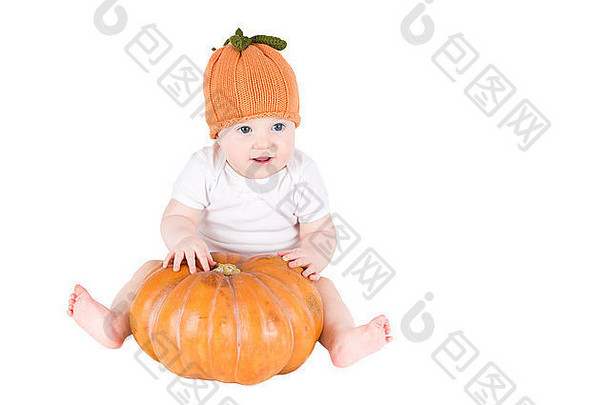 有趣的婴儿坐在一个南瓜旁边，戴着一顶编织的南瓜帽，隔离在白色的地板上