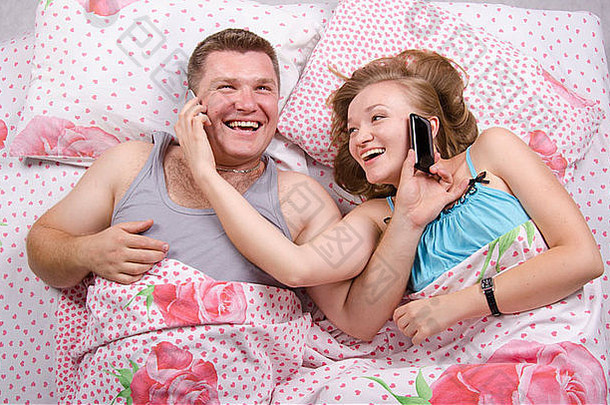 躺在床上的年轻夫妇。每个人都在我的手机里。与打电话时的愉快和喜悦搭配。照片制作于顶部