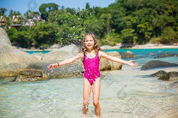 可爱快乐的小女孩穿着游泳衣沿着海滩奔跑，跳过海浪。美丽的夏日阳光明媚，碧海碧海，岩石嶙峋。
