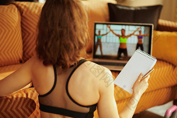 从后面可以看到一位穿着健身服的年轻女子，她拿着记事本和笔，通过笔记本电脑在网上观看健身视频