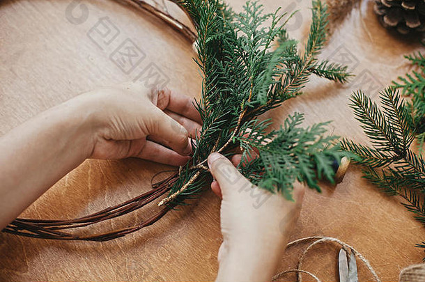 手拿着雪松树枝和松果，线，剪刀放在木桌上。制作圣诞花环车间的详细信息。制作质朴的圣诞节