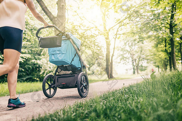 带着婴儿车的跑步妇女在公园里享受夏日。慢跑或电动步行超级妈妈，带婴儿慢跑的活跃家庭。