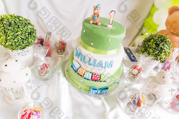 白色和绿色的生日蛋糕，白色桌子上放着一岁的蜡烛和泰迪熊