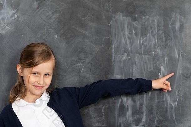 一个女孩微笑着站在黑板前，指着一块空白处，准备用粉笔写字。