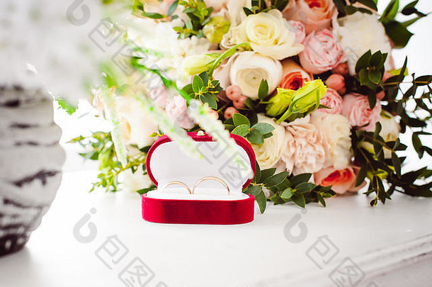 一个红色天鹅绒盒子的照片，盒子上有新娘和新郎的结婚戒指，在一张白色的桌子上，盒子上有一束新娘花