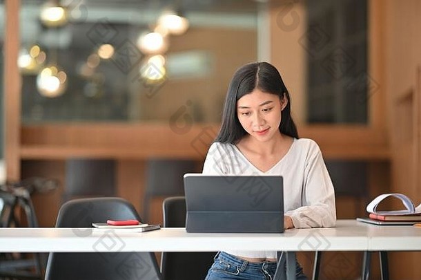 年轻漂亮的女士坐在舒适的工作台上，以现代客厅为背景，通过平板电脑上网。