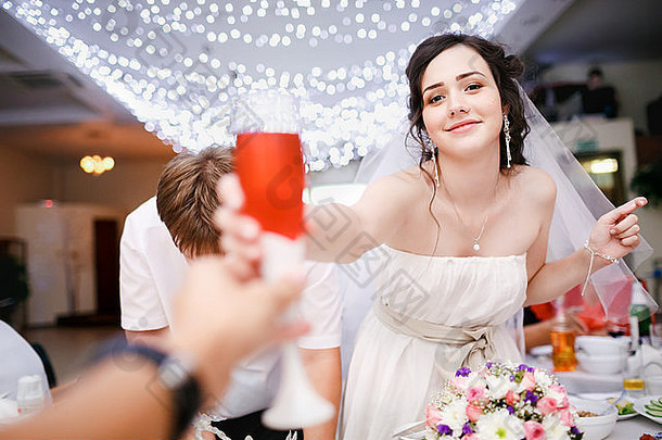 身着白色礼服的年轻美丽新娘开玩笑地举起一杯香槟，想干杯，碰杯。餐厅婚礼宴会，天花板花环。庆祝。干杯