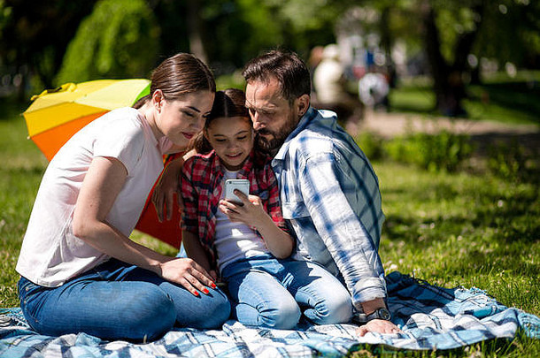 一家人坐在公园的毯子上，微笑着用手机看照片。
