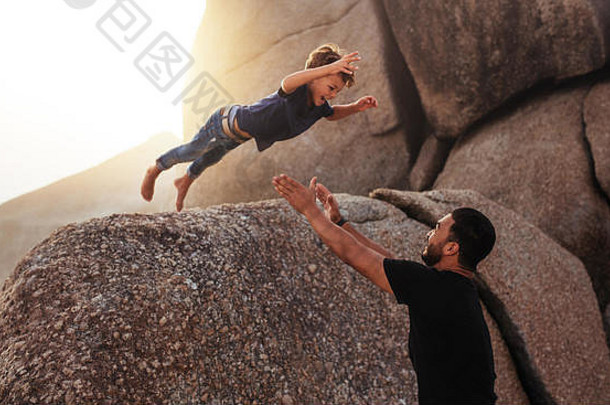 一张可爱的小男孩从一块大石头上跳到他父亲怀里的照片。父亲和儿子在海滩上享受暑假。