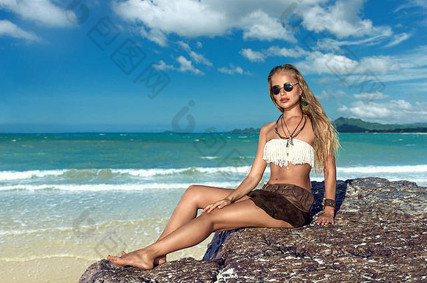 热带海滩上坐在石头上的年轻时尚女郎