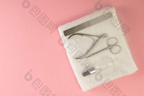 指甲锯、角质层剪、指甲剪和手油放在粉色背景上的白色毛巾上。俯视图