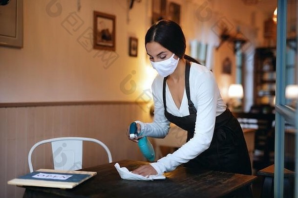 戴着口罩的年轻女子在咖啡馆的室内工作，为桌子消毒。