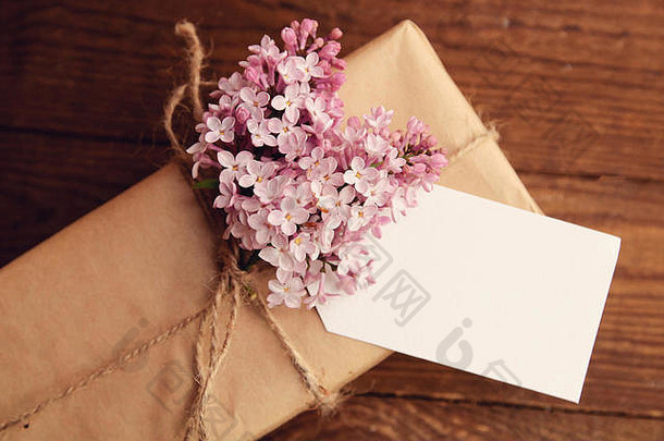 用<strong>牛皮纸包装</strong>的礼物放在木桌上，用绳子绑着，还有一束粉红色的丁香花，外加贺卡