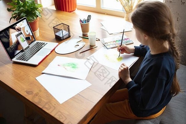 女孩在家里用颜料和铅笔画画，在笔记本电脑上看老师的在线教程。数字化、远程教育理念。技术、设备。男子表演，在线授课。艺术品