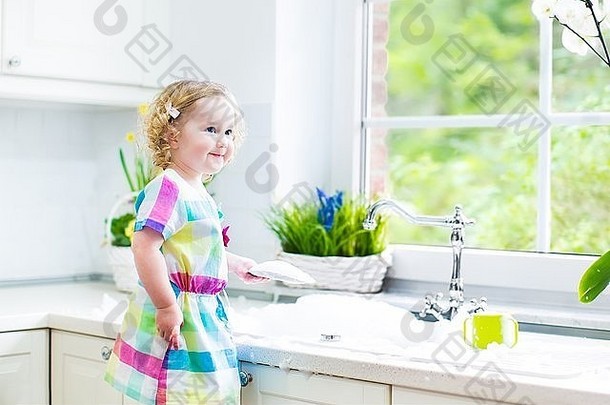 可爱的卷曲的蹒跚学步的小女孩穿着五颜六色的衣服洗盘子，用海绵清洁，在水槽里玩泡沫