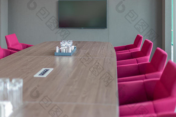 一张棕色的会议桌被红色扶手椅包围，背景墙上有一台电视