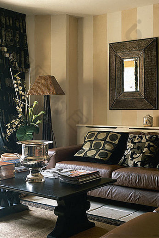 客厅的棕色真皮沙发上方有厚重的木框镜子，沙发上有调色垫，还有咖啡桌和条纹墙纸