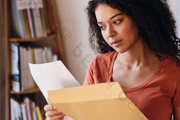 一位年轻严肃的女士，深色卷发，穿着T恤衫，若有所思地看着家里手里拿着考试成绩的信