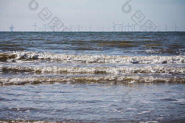 英国北威尔士普雷斯塔廷中央海滩的潮水滚滚而来。背景中的一排风力涡轮机（风电场）在地平线上。