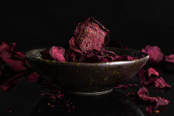 很多片干红色的甜菜根周围休息黑暗陶瓷碗孤立的黑色的玻璃