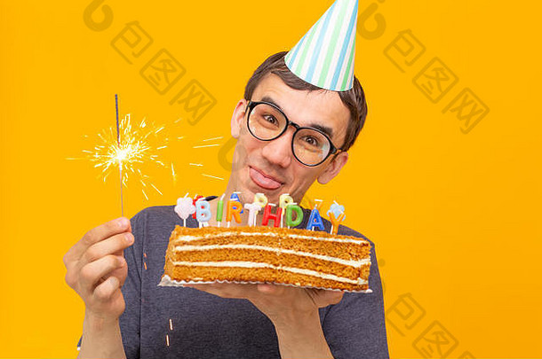 一个戴着眼镜、戴着纸礼帽、手持蛋糕、站在黄色背景上的疯狂快乐的年轻人，生日快乐。周年庆祝