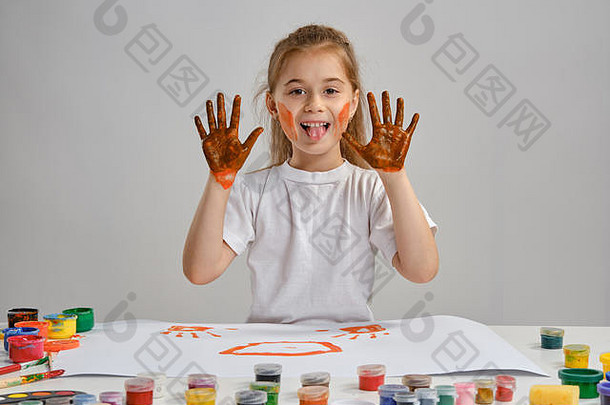 穿着白色t恤的小女孩坐在桌旁，手里拿着惠特曼和五颜六色的颜料，展示着她画过的手。白色的隔离带。中等特写镜头。