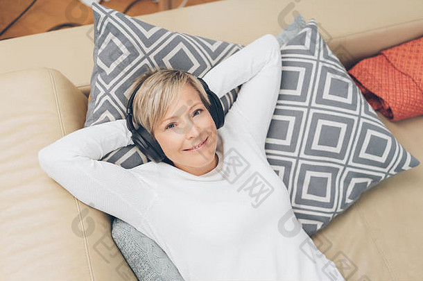 幸福的女人躺在沙发上对着摄像机微笑，在家里放松，戴着立体声耳机听音乐