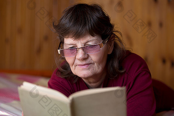 戴着眼镜的可爱老妇人在看书，舒适地躺在家里的床上，在一间有着木制墙壁的舒适的乡村房间里，享受着爱伦·坡