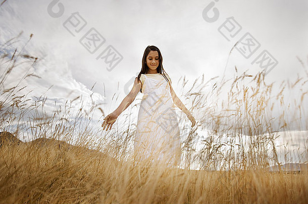 一个穿着白色衣服的女孩在长草地上，两臂伸出在身体两侧向下看。