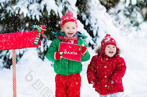 圣诞树下雪地里的红色信箱里，戴着驯鹿编织帽和围巾的快乐的孩子们手里拿着给圣诞老人的信和圣诞礼物心愿单