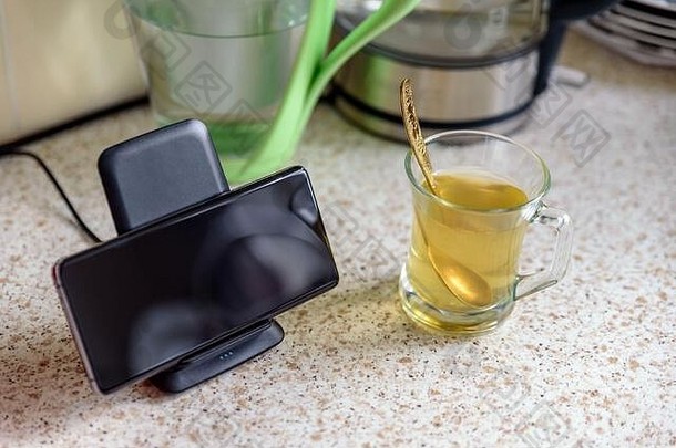 智能手机无线充电在厨房桌面茶杯旁边的充电座上。
