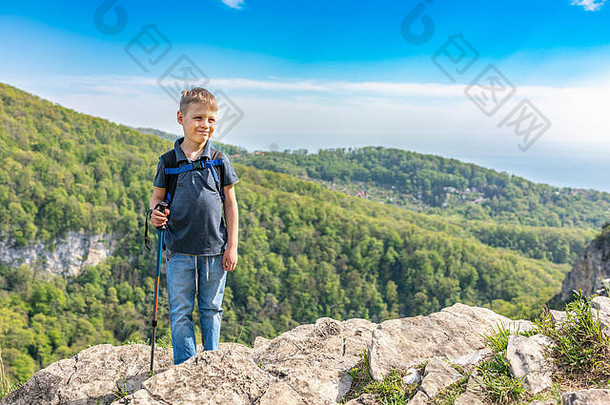 一个带着登山杖和背包微笑的男孩站在绿色森林中的山顶上。山上春夏晴朗