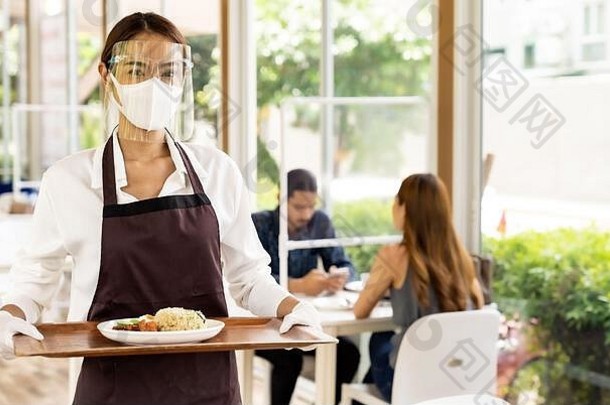 肖像迷人的亚洲女服务员戴着口罩和面罩，端着餐盘，以顾客为背景为顾客端饭。新常态