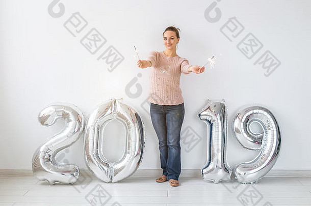 新的2019年即将到来。在白色的房间里，站在银色数字和火花旁边的快乐的年轻女子