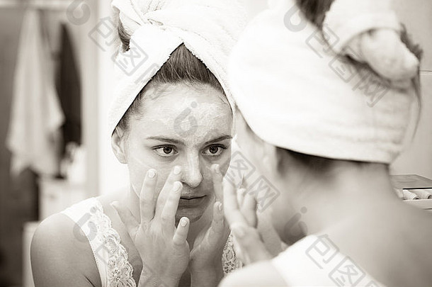女人在脸上涂面膜保湿护肤霜，对着浴室镜子看。照顾自己肤色的女孩。