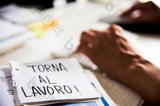 一名男子坐在办公桌旁的特写镜头，前景中有一张便条，上面写着“torna al-loro”，背面用意大利语写着“回到工作岗位”