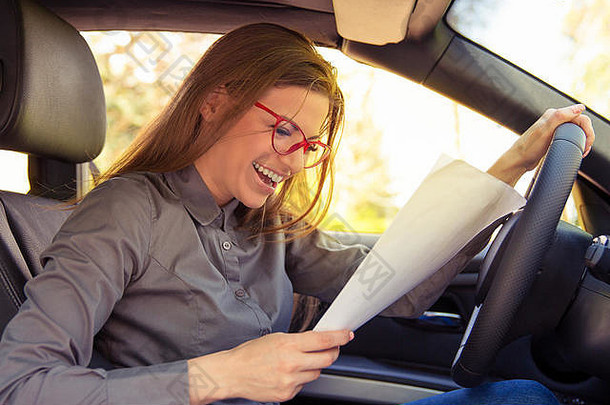 戴眼镜的年轻女子侧视图，她坐在车里愉快地微笑着，一边阅读<strong>试卷</strong>和测试结果