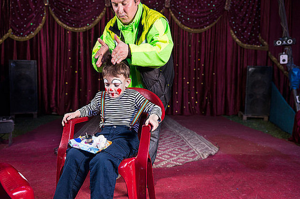 小男孩扮成小丑坐在椅子上，化妆盘放在舞台上的大腿上，男人穿着鲜艳的夹克，双手放在男孩的头顶上。