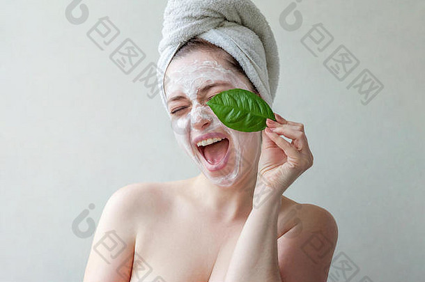 一位面带微笑的黑发女子的美丽肖像，头戴毛巾，脸上涂有白色滋养面膜或乳霜，手拿绿叶，背景为白色。护肤清洁生态有机化妆品spa放松概念