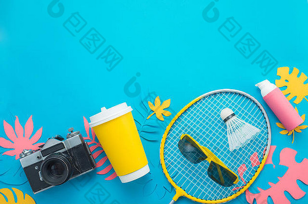 夏季活动标题。羽毛球火箭与热带树叶，人字拖，太阳镜，防晒霜，相机和纸杯从上方明亮的蓝色b