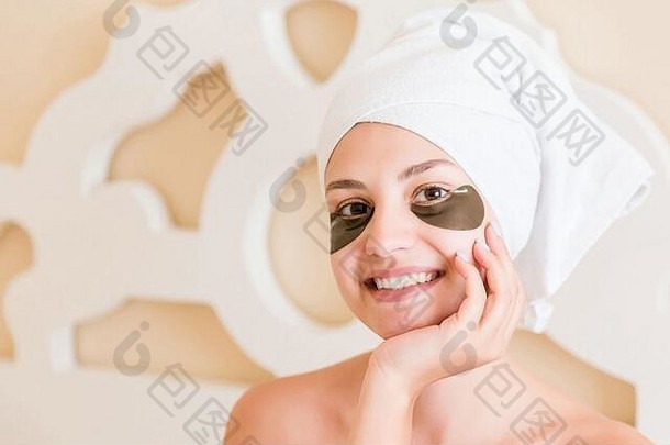 沐浴后，一位面带微笑的妇女裹着毛巾，眼睛下面有黑色的斑点，她在床上摆姿势。美容概念。