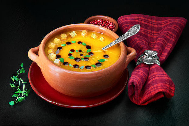 南瓜奶油汤装在陶瓷碗中，配以南瓜油、百里香叶和面包片，餐巾以深色石头为背景，周围有香料