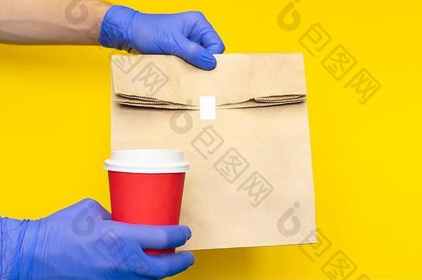 快递员戴着医用手套，手持纸袋午餐，咖啡杯隔离在黄色背景上。检疫科罗纳病毒中的非接触式递送服务