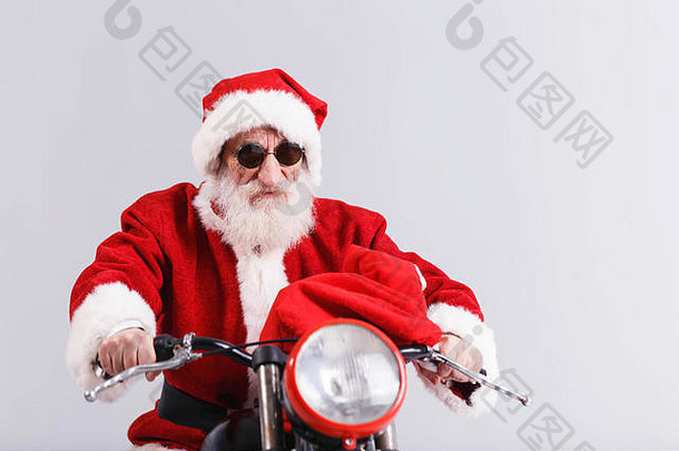 带着白胡子的圣诞老人戴着太阳镜，穿着圣诞套装骑着摩托车，包里装着礼物、新年、圣诞节、节日、纪念品、礼品、<strong>购</strong>物、折扣、商店、雪人圣诞老人、化妆、发型、<strong>狂欢</strong>节。