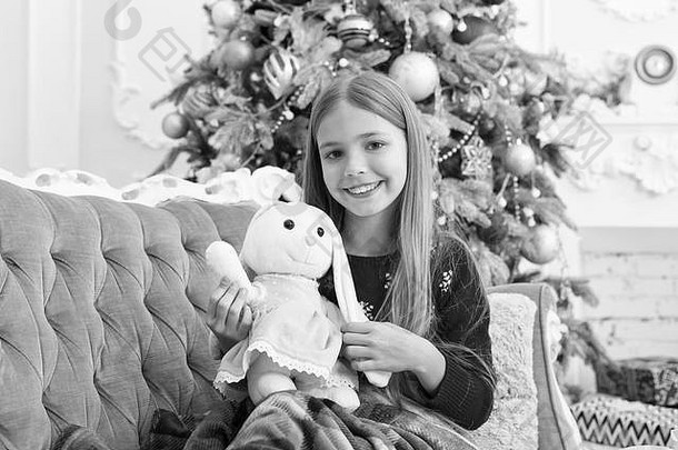 最好的圣诞玩具。小女孩和可爱的兔子在圣诞树旁。小女孩抱着兔子玩具。小孩玩软玩具。小孩带着礼物开心地笑着。最令人期待的玩具。