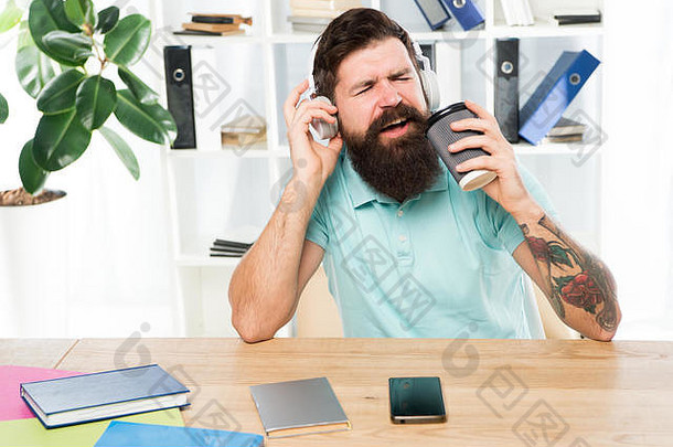 正常工作日。留胡子<strong>的</strong>男人戴着耳机坐在办公室听音乐唱歌。拿着咖啡杯<strong>的</strong>工人用最喜欢<strong>的歌曲</strong>开始工作日。充满灵感和活力。办公室生活<strong>节奏</strong>。