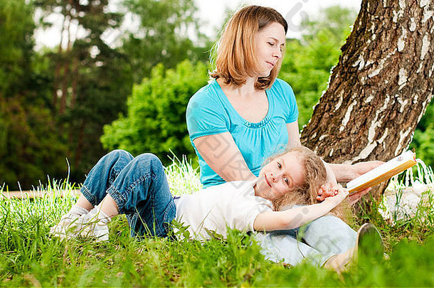 年轻的母亲坐在<strong>树下</strong>的草地上，给躺在母亲膝上微笑着的小女儿<strong>读书</strong>