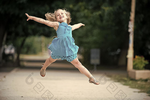 小或可爱快乐的孩子，带着可爱的笑脸，身着蓝色连衣裙，金发鞠躬，在夏日户外的模糊背景下跳跃。