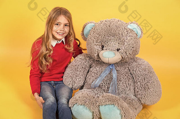 孩子带着灰色的软玩具微笑。女孩和橙色背景的大泰迪熊坐在一起。带动物娃娃的孩子，礼物和礼物。节日、生日、<strong>周年庆</strong>典。快乐童年的概念。